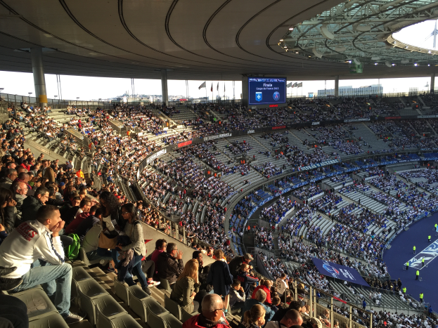 Eine große Menschenmenge sitzt in einem Stadion und schaut ein Fußballspiel, mit einer Bühne auf der rechten Seite, Fahnen, Stangen und einem Bildschirm im Hintergrund, unter einem sichtbaren Himmel, im Allianz Arena in München, Deutschland.