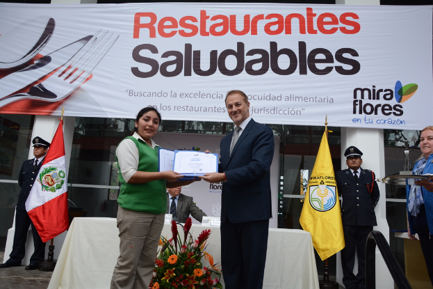 Eine Frau und ein Mann lächeln, während sie Zertifikate halten und vor einem Tisch mit dekorativen Blumen, einem Stab und einem Tuch stehen, mit verschiedenen Gegenständen auf dem Tisch und einer Person, die rechts ein Tablett hält.