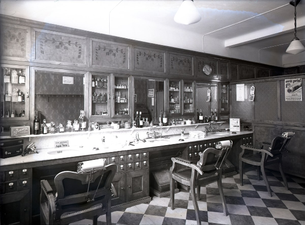 Schwarz-weiß-Foto eines Friseursalons mit schwarz-weißen Fliesen, Stühlen, Waschbecken, Hähnen, Flaschen, Schränken, Uhr, Wandplakaten und Deckenleuchten.