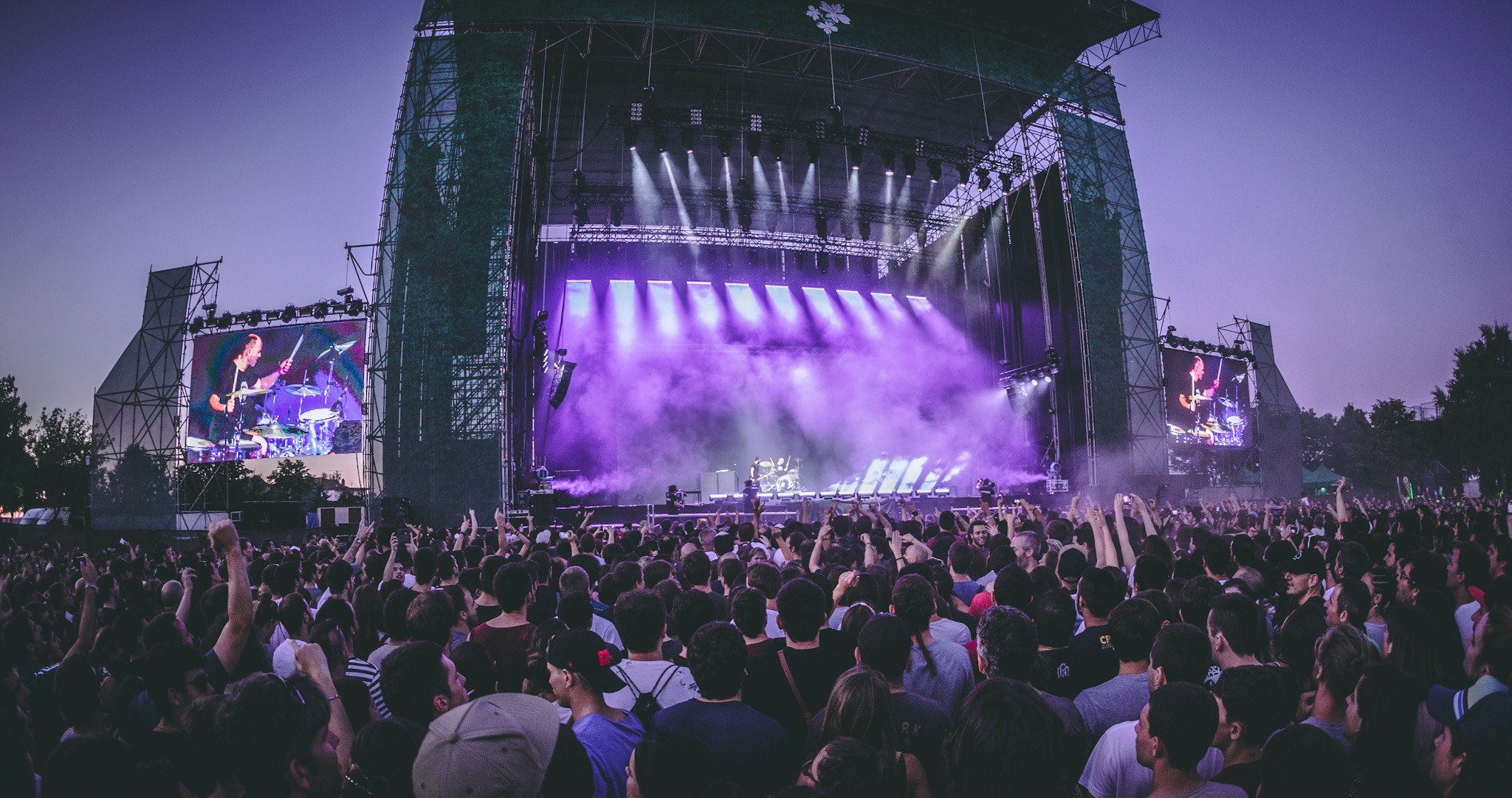 Eine große Menschenmenge auf einem Musikfestival mit einer Bühne, auf der Musiker Instrumente spielen, beleuchtet von Lichtern und umgeben von Bildschirmen, vor einer Kulisse aus einem Gebäude, Bäumen und einem klaren blauen Himmel.