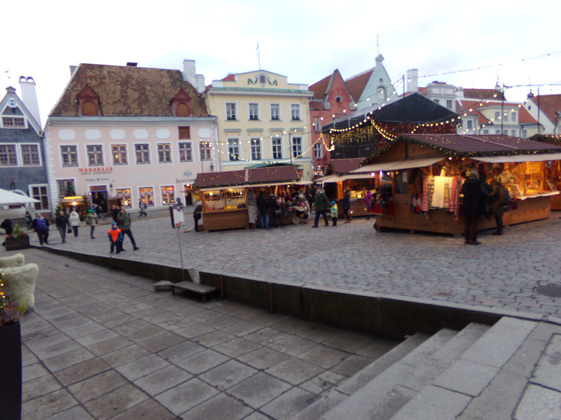 Ein lebendiger Weihnachtsmarkt in Tallinn, Estland, mit Menschen um beleuchtete und dekorierte Stände, Gebäuden mit Fenstern im Hintergrund, bewölktem Himmel, Treppen, die zum Markt führen, und Töpfen mit Pflanzen, die herumstehen.