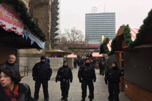 Polizisten in schwarzen Uniformen und Mützen vor einem geschmückten Weihnachtsmarkt in Berlin, mit festlichen Dekorationen, Gebäuden, Bäumen und einem klaren blauen Himmel im Hintergrund.