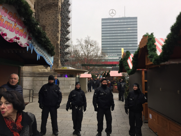 Polizisten in schwarzen Uniformen und Mützen vor einem geschmückten Weihnachtsmarkt in Berlin, mit festlichen Dekorationen, Gebäuden, Bäumen und einem klaren blauen Himmel im Hintergrund.