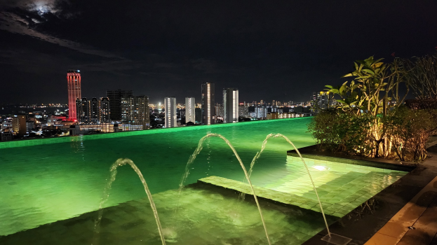 Ein Schwimmbad mit einem zentralen Brunnen, umgeben von Pflanzen auf der rechten Seite und beleuchteten Gebäuden im Hintergrund bei Nacht.