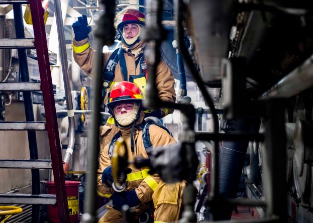 Zwei Feuerwehrleute in Helmen und Handschuhen steigen eine Leiter in einem Gebäude hoch, mit Rohren und anderen Gegenständen auf der rechten Seite.