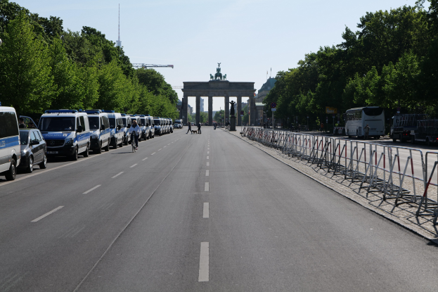 Lange Reihe von Polizeiwagen auf der Straße vor dem Brandenburger Tor in Berlin, Deutschland, mit Menschen, die Fahrräder fahren und auf der Straße stehen, Barrieren, Bäume, ein Tor mit Statuen im Hintergrund und sichtbarer Himmel.