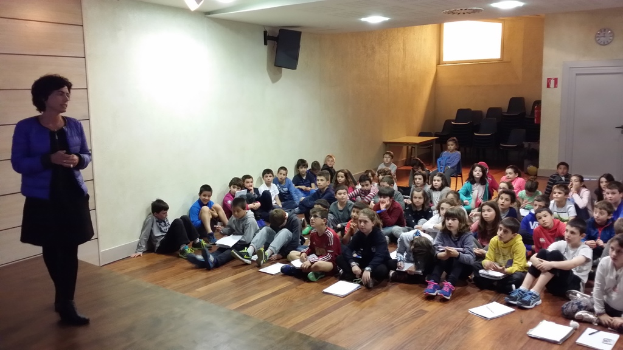 Eine Gruppe von Kindern, Jungen und Mädchen, sitzt auf dem Boden mit Büchern vor sich, während eine Frau auf der Bühne links steht, mit einer Wand im Hintergrund.