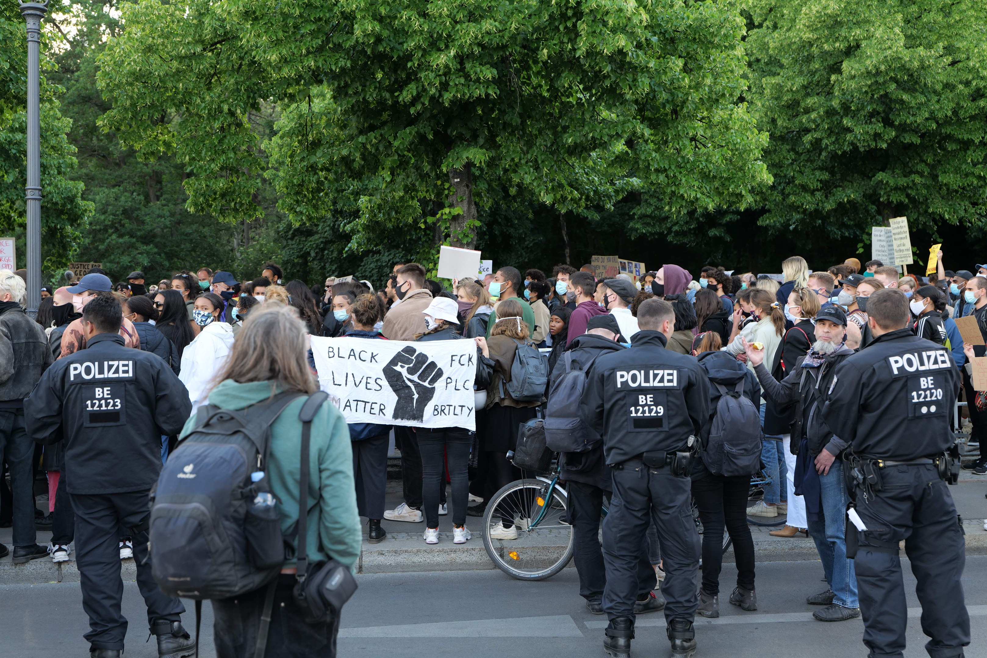 Eine große Gruppe von Menschen nimmt an einer Black Lives Matter Demonstration in Berlin teil, einige halten Schilder und andere tragen Mützen und Taschen, vorne steht ein Fahrrad und im Hintergrund sind Bäume und ein Pfahl zu sehen.