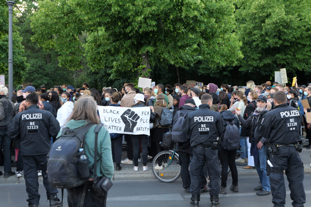 Eine große Gruppe von Menschen nimmt an einer Black Lives Matter Demonstration in Berlin teil, einige halten Schilder und andere tragen Mützen und Taschen, vorne steht ein Fahrrad und im Hintergrund sind Bäume und ein Pfahl zu sehen.