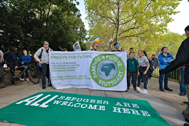 Eine Gruppe von Menschen steht auf dem Boden, einige halten ein Banner mit der Aufschrift "All Refugees Are Welcome Here" und eine Flagge, mit Fahrrädern, einem Zaun, einem Straßenschild, einer Tafel, Bäumen und einem bewölkten Himmel im Hintergrund.
