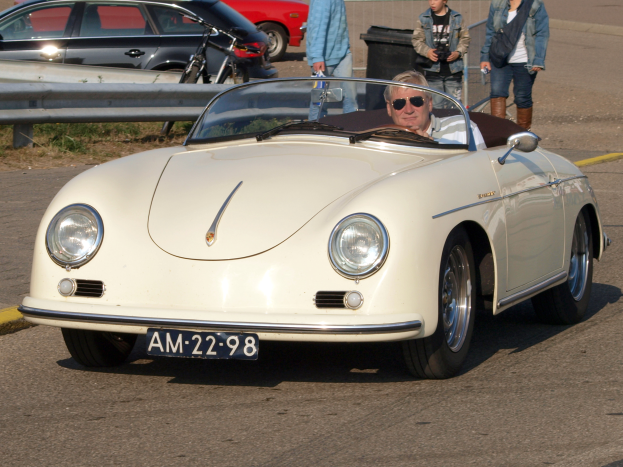 Ein Mann fährt einen weißer Porsche 356 Speedster die Straße entlang, umgeben von Menschen in verschiedenen farbigen Kleidern, mit einem Geländer und einem schwarzen Mülleimer an der Seite und weiteren Autos und einem Gebäude im Hintergrund.