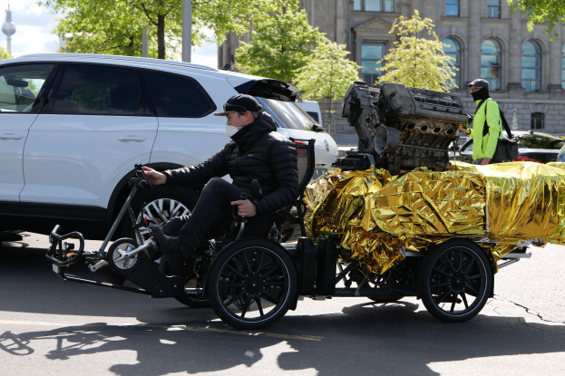 Ein Mann im Rollstuhl mit einem großen Motor daran befestigt, umgeben von Fahrzeugen auf einer Straße mit Bäumen, Gebäuden und einem klaren blauen Himmel im Hintergrund.