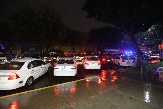 Eine Gruppe von Polizeiwagen auf einem Parkplatz bei Nacht geparkt, mit einem Schild, Gras, Bäumen, Gebäuden, einer Fahne, Lichtern und einem bewölkten Himmel.