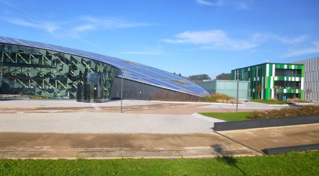 Die Universität der Künste und Wissenschaften in Frankreich, ein großes Gebäude mit einem grünen Dach, befindet sich in einem Park umgeben von Gras, Pflanzen, Blumen, Pfählen und Bäumen, mit einem Himmel voller weißer, flauschiger Wolken im Hintergrund.