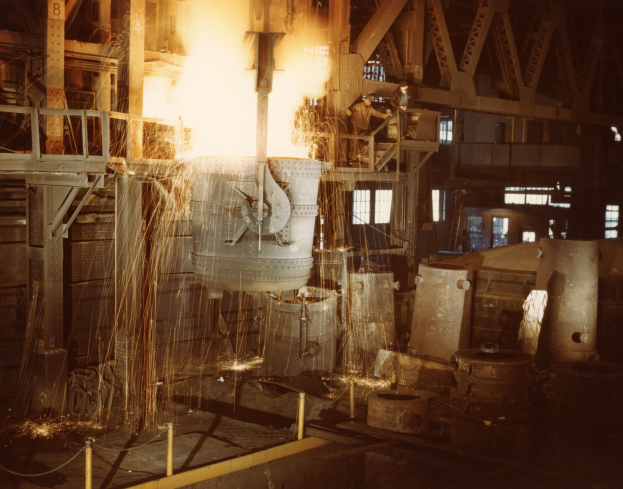 Großes Metallobjekt wird in einen Ofen gegossen, umgeben von Menschen, beleuchtet vom Feuer des Ofens, in einer Fabrikumgebung mit Metallobjekten, Polen und Fenstern im Hintergrund.