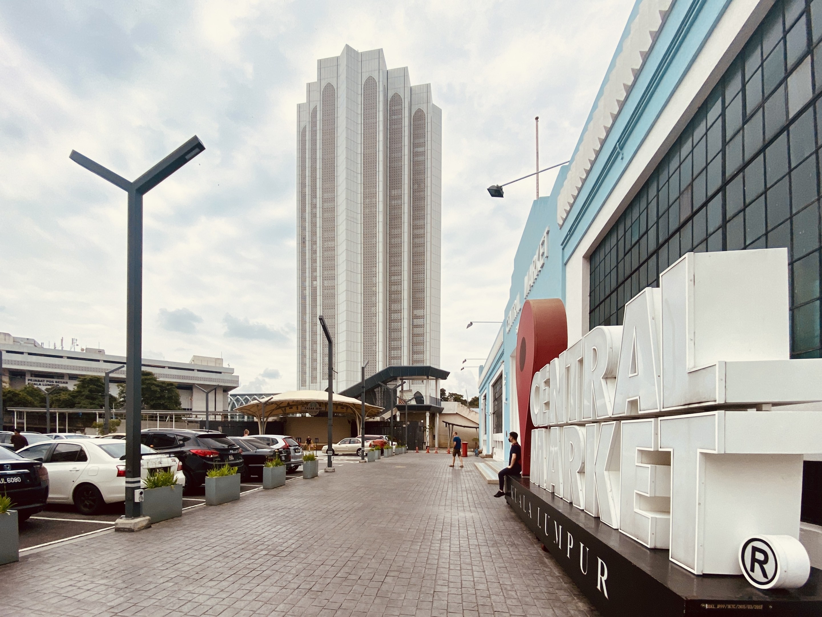 Ein belebter zentraler Markt in Kuala Lumpur, Malaysia, mit hohen Gebäuden, Straßenmasten, Straßenlaternen, Kraftfahrzeugen, Fußgängern, Namensschildern, Bäumen und einem bewölkten Himmel.