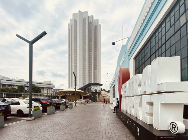 Ein belebter zentraler Markt in Kuala Lumpur, Malaysia, mit hohen Gebäuden, Straßenmasten, Straßenlaternen, Kraftfahrzeugen, Fußgängern, Namensschildern, Bäumen und einem bewölkten Himmel.