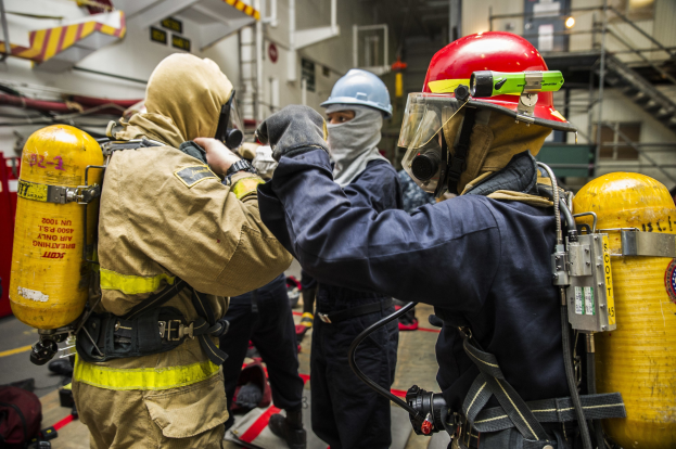 Feuerwehrleute in Schutzausrüstung, einschließlich Helmen, Handschuhen und Sauerstoffzylindern, stehen zusammen mit Equipment-Bags auf dem Boden in der Nähe von Treppen und Geländern.