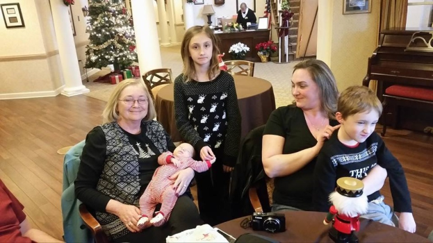 Ein Raum mit einer Wand, die ein Fotodisplay, einen Weihnachtsbaum, eine Lampe und Pflanzen mit Blumen zeigt. Es gibt Treppen, einige Menschen, die auf Stühlen und Tischen sitzen, mit einem Spielzeug und einer Kamera auf einem der Tische.