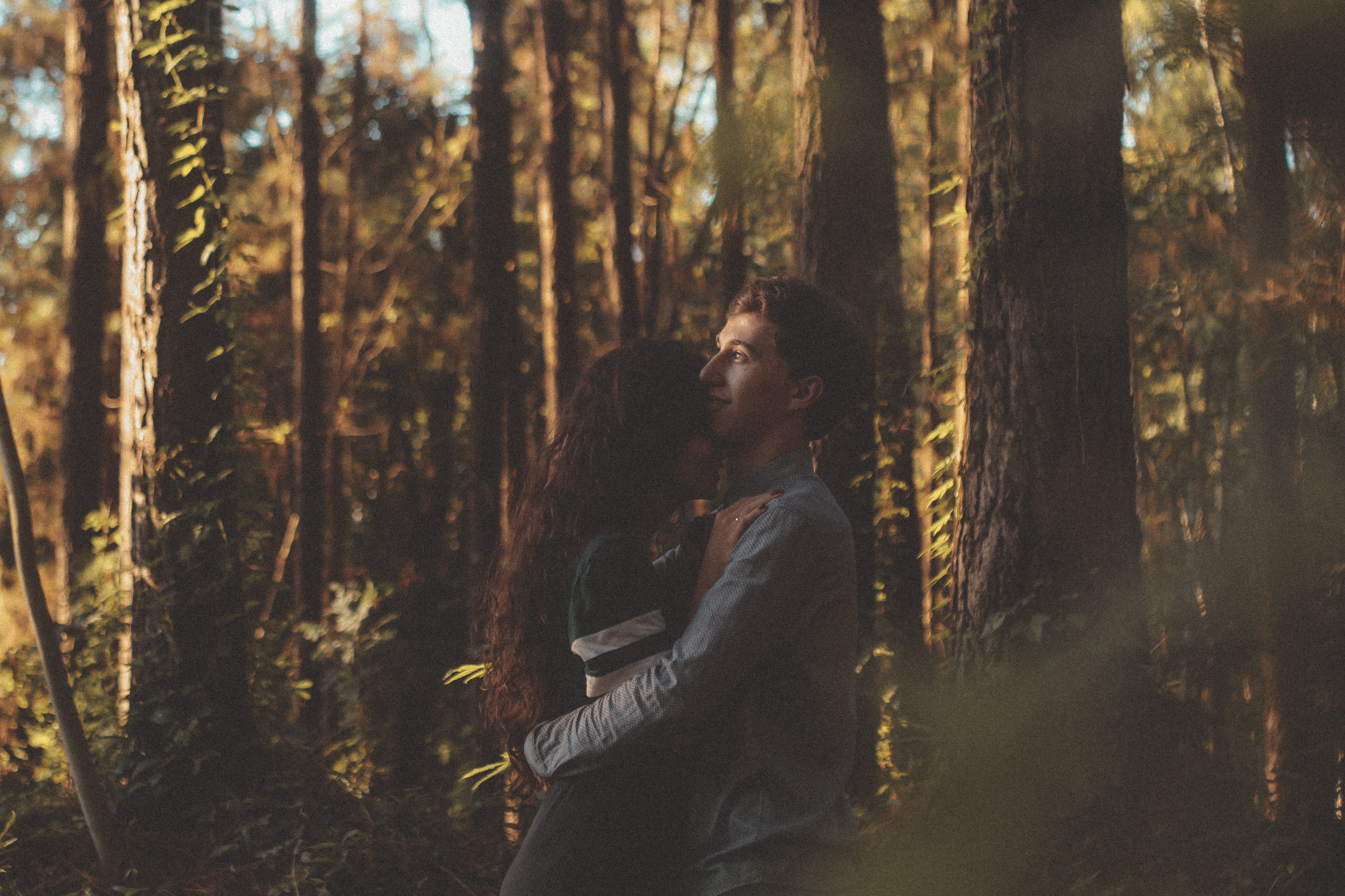 Ein Mann und eine Frau umarmen und küssen sich in einem bewaldeten Gebiet mit hohen Bäumen und üppiger Vegetation unter einem sichtbaren Himmel während ihrer Verlobungsfotoshoot.
