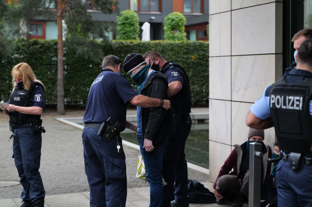 Eine Gruppe von Polizisten steht vor einem Gebäude, eine Frau hält ein Buch und einen Stift in der Hand; Menschen stehen und sitzen auf dem Boden, und Pflanzen, Bäume und Gebäude sind im Hintergrund zu sehen.