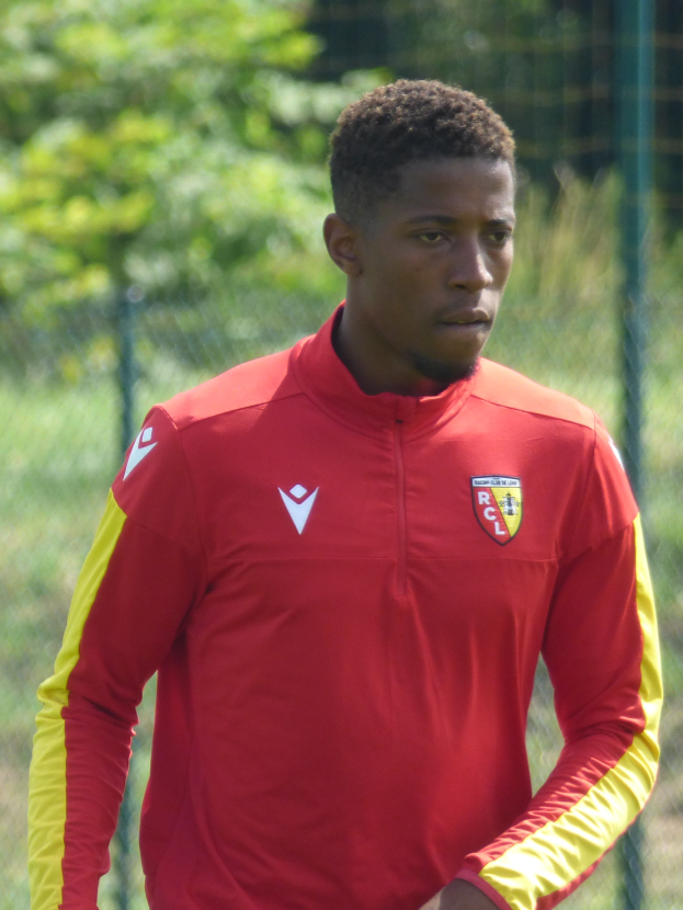 Ein junger Mann in einem roten und gelben Trikot steht auf einem Fußballfeld umgeben von einem Zaun und Bäumen und sieht entschlossen aus.