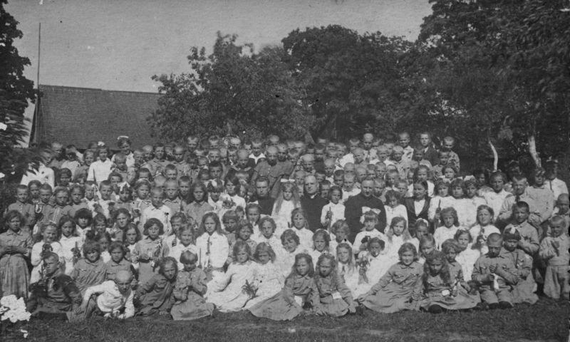 Schwarz-weiß-Foto einer Gruppe von Kindern, die vor einem Haus mit Bäumen und einem klaren Himmel stehen.