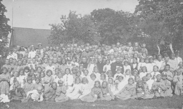 Schwarz-weiß-Foto einer Gruppe von Kindern, die vor einem Haus mit Bäumen und einem klaren Himmel stehen.