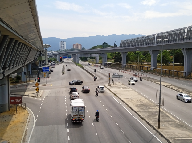 Eine belebte Autobahn mit Verkehr, neben einer Brücke, mit Straßenlaternen, Verkehrssignalen, Schildern, Bäumen, Gebäuden, Höhen und einem bewölktem Himmel im Hintergrund.