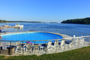 Ein großes Schwimmbad mit Liegestühlen und einem Whirlpool auf einer grasbewachsenen Uferpromenade, die von einem Geländer umgeben ist, mit Booten auf einem See, Bäumen und einem klaren blauen Himmel im Hintergrund.