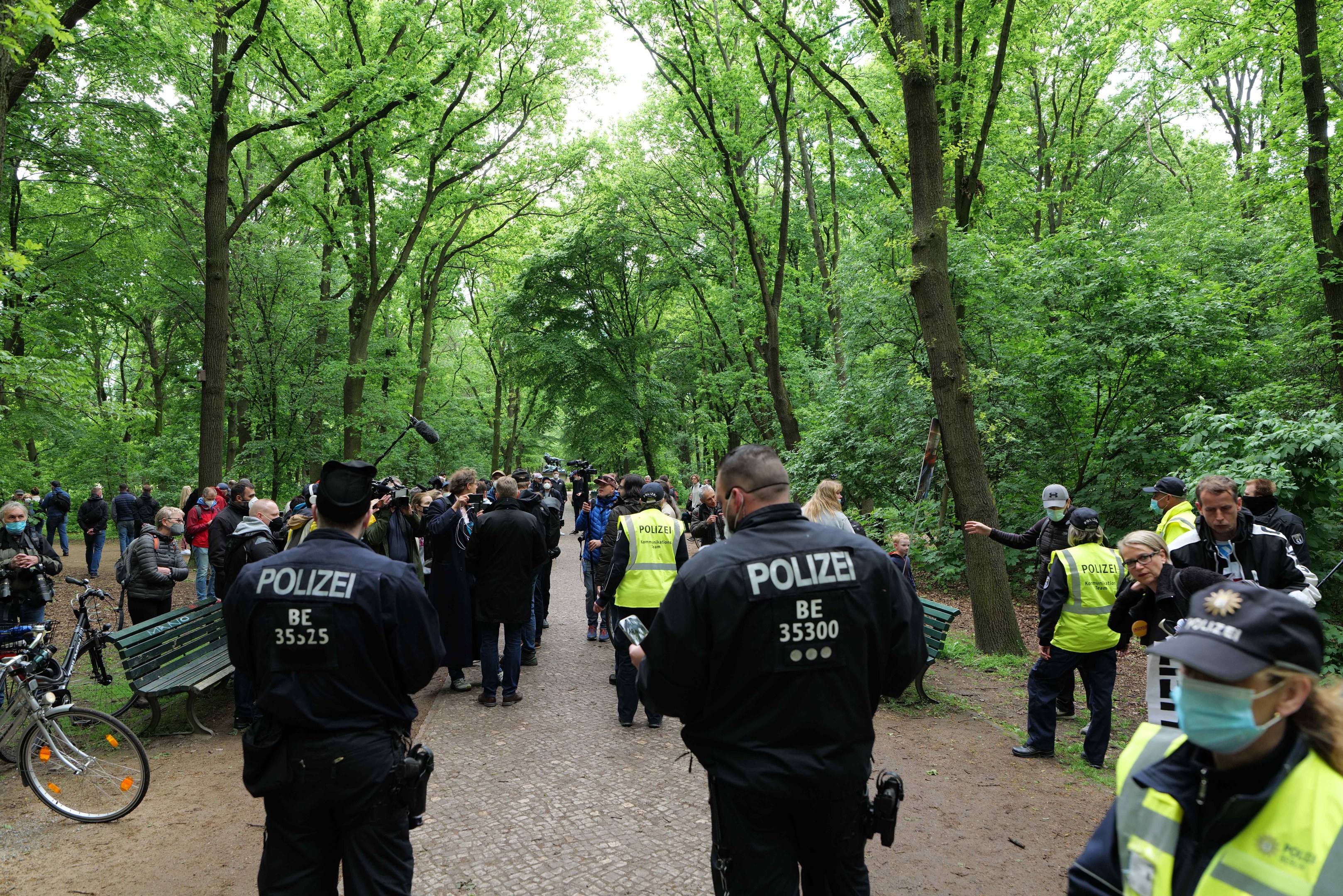 Polizisten vor einer Menschenmenge bei einer Outdoor-Veranstaltung mit Fahrrädern und einer Bank im Vordergrund und Bäumen im Hintergrund