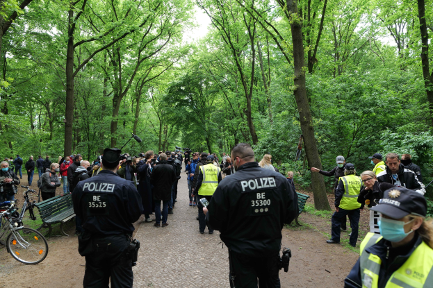 Polizisten vor einer Menschenmenge bei einer Outdoor-Veranstaltung mit Fahrrädern und einer Bank im Vordergrund und Bäumen im Hintergrund