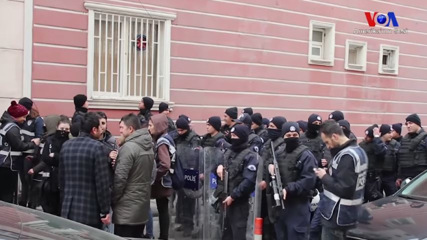 Eine Gruppe von Menschen, einige bewaffnet und mit Mützen, steht vor einem Gebäude mit Fenstern und hat sich versammelt, um gegen eine Regierungsentscheidung zu protestieren, mit Fahrzeugen und einem Verkehrskegel im Vordergrund und einem Wasserzeichen auf dem Bild.