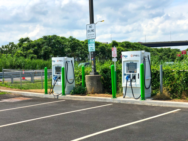 Elektrofahrzeug-Ladestation auf einem Parkplatz mit umgebender Infrastruktur, Vegetation und einer Brücke im Hintergrund mit Wolkenhimmel.