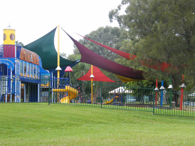 Ein Park mit einem eingezäunten Spielplatz, der eine Rutsche, Stangen und ein Zelt enthält und von grünem Gras, Bäumen und einem klaren blauen Himmel umgeben ist, wo Kinder spielen.