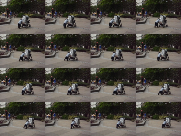 Ein Kind geht auf einer Straße und zieht einen Wagen, mit zwei Personen auf der linken Seite, die Fahrräder halten, Bäume im Hintergrund und ein Gebäude auf der linken Seite.