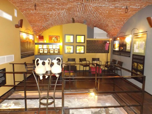 Ein Museumsraum voller Tische, Stühle, ein Geländer, Vasen, Texttafeln, Wandrahmen, Lampen und verschiedene Ausstellungsstücke.