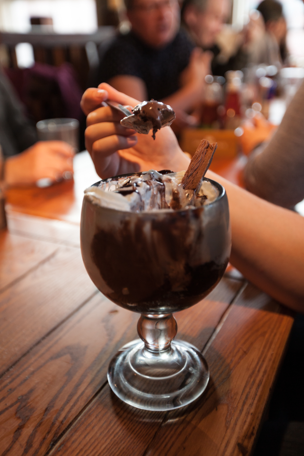 Eine Gruppe von Menschen sitzt um einen Tisch mit einem Glas Schokoladenpudding in der Mitte, eine Person im Vordergrund hält einen Löffel und isst den Pudding, mit einem unscharfen Hintergrund.