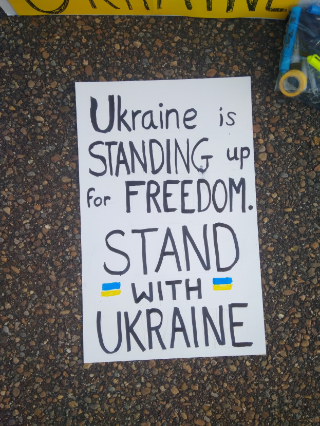 Ein weißer Zettel mit der Aufschrift "Ukraine steht für Freiheit auf, stellt Euch mit Ukraine" liegt vor einer gelben Fahne auf dem Boden. Rechts daneben befindet sich eine Plastikhülle mit verschiedenen Gegenständen.