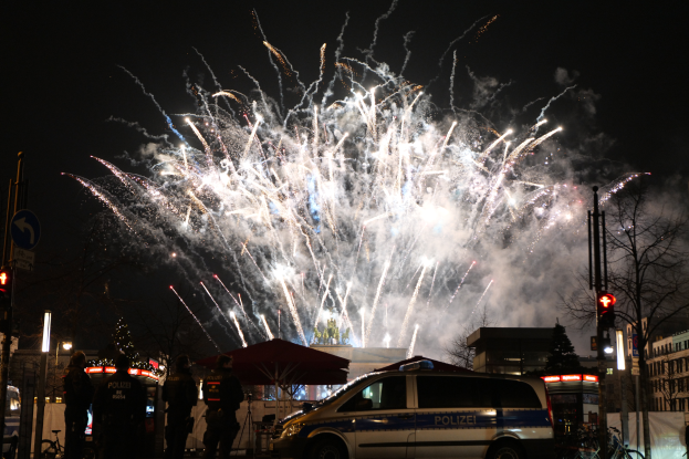 Ein Polizeiwagen steht vor einer Menschenmenge, mit beleuchteten Gebäuden und Bäumen im Hintergrund, während farbige Feuerwerke den Himmel erhellen und das Neujahrsfest in Berlin feiern.