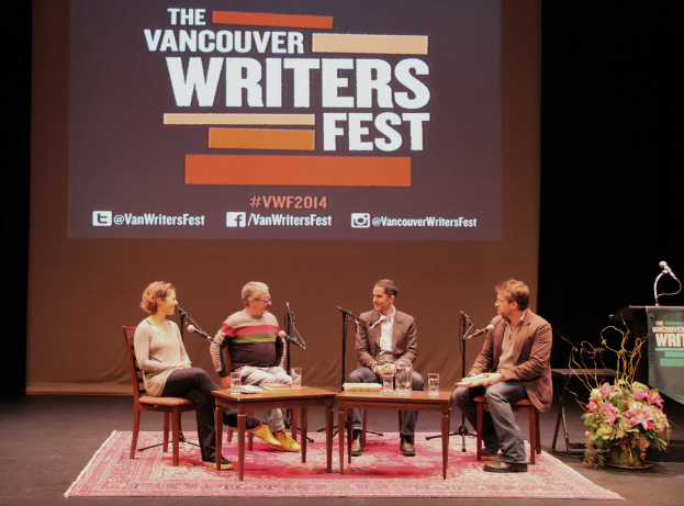 Eine Gruppe von Männern und einer Frau, die auf Stühlen auf einer Bühne sitzen und in Mikrofone sprechen, mit einem Bildschirm im Hintergrund, einem Teppich auf dem Boden, einem Tisch in der Mitte und einem Blumentopf in der Nähe.