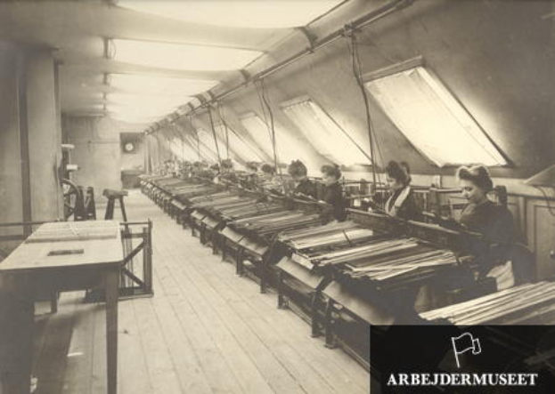 Ein Schwarz-Weiß-Foto von Frauen in einer Textilfabrik, die an Maschinen mit Tischen auf der linken Seite sitzen, eine Uhr an der Wand im Hintergrund und Text in der rechten unteren Ecke.