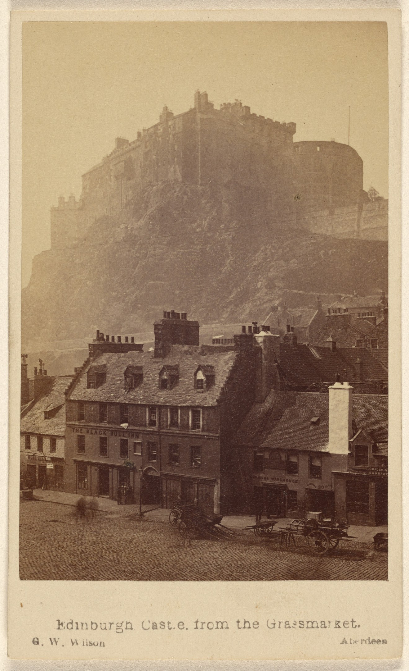 Altes Foto von Edinburgh Castle aus der Sicht vom Grassmarket, mit Gebäuden und Karren im Vordergrund und einem Hügel im Hintergrund; Text ist am unteren Bildrand sichtbar.