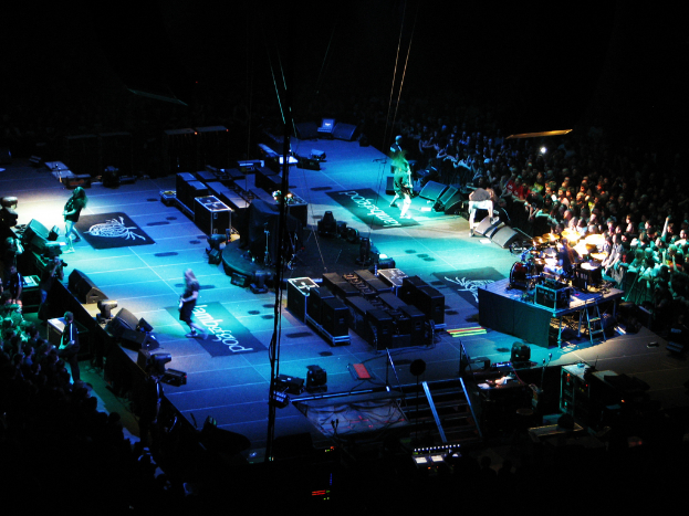 Leute, die auf einer Bühne mit Kisten, Lautsprechern, Lichtern und Mikrofonen performen, mit einer großen Menge auf beiden Seiten und einem dunklen Hintergrund.