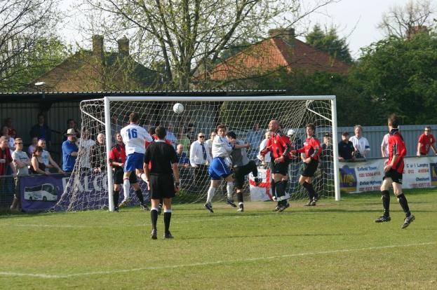 Fußballspieler bei einem Spiel auf einem Feld mit einem Tor, während Zuschauer dahinter stehen, mit Bäumen und Häusern im Hintergrund.