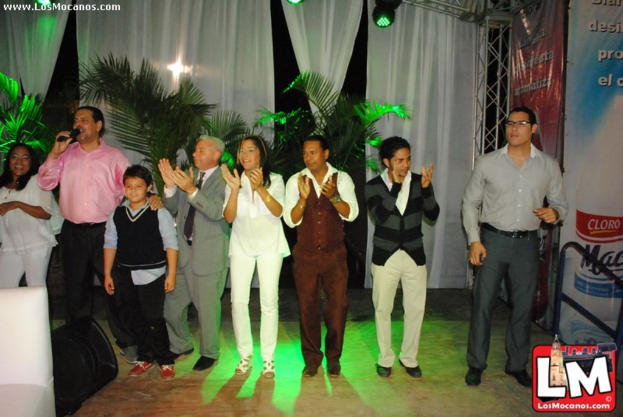 Eine Gruppe von Menschen auf einer Bühne mit einigen applaudierenden Personen, einer Person mit Mikrofon, Hosen und Vorhängen dahinter und Lampen oben.