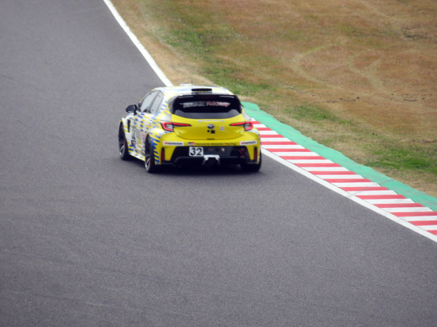 Ein gelbes Sportauto mit Text und Zahlen darauf, das eine Rennstrecke entlangfährt, die von Gras gesäumt ist.