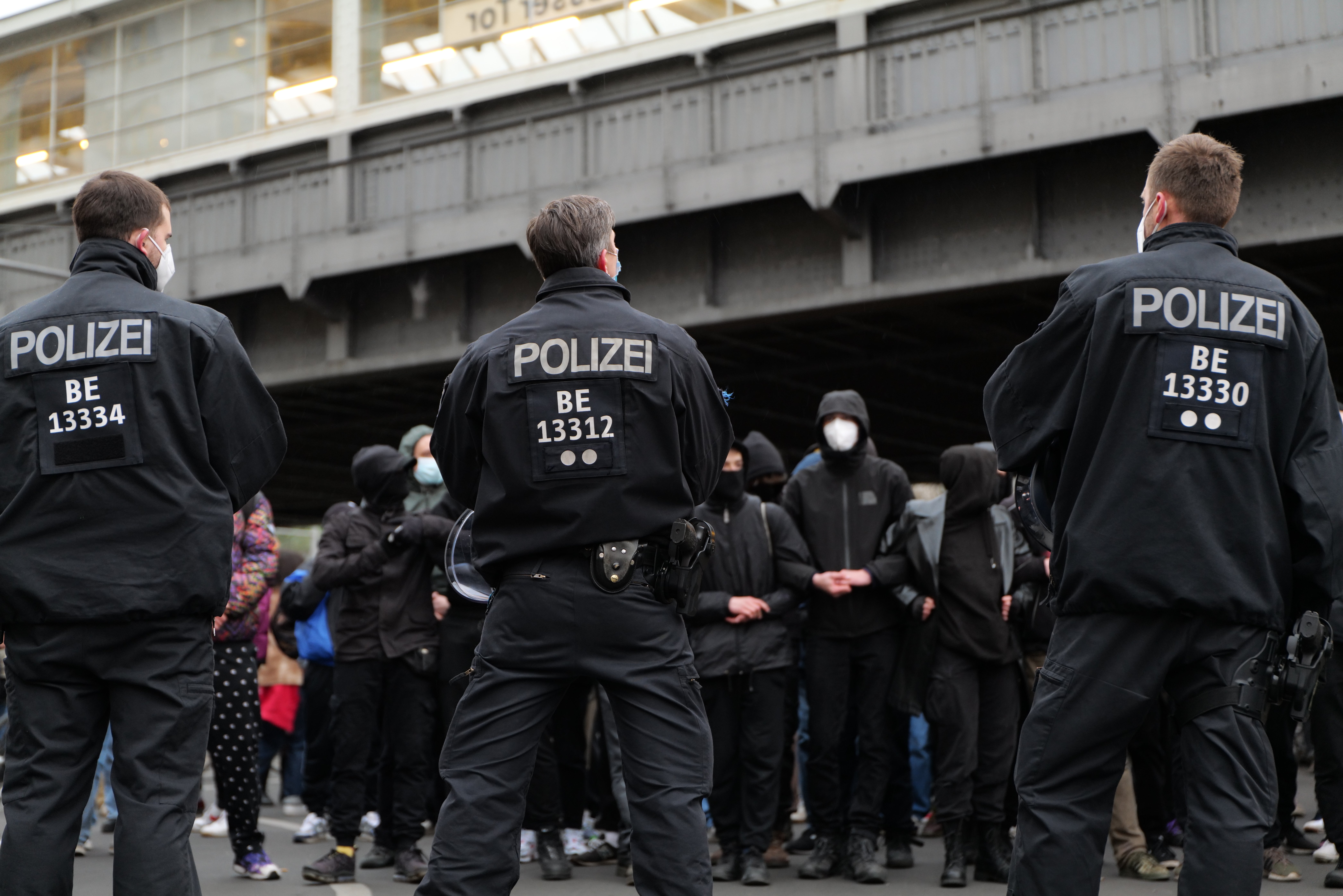 Eine Gruppe uniformierter Polizisten steht vor einer Menge von Menschen in schwarzen Uniformen und Masken, im Hintergrund eine Brücke und ein Gebäude, was auf eine städtische Umgebung hindeutet.