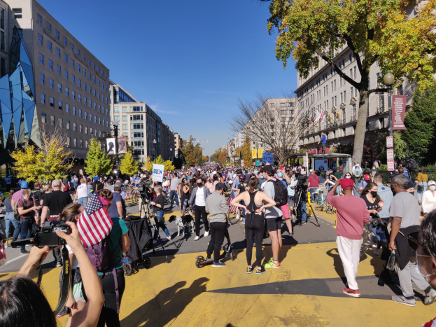 Eine große Gruppe von Menschen, die eine Straße in Washington, DC, entlanggehen, gesäumt von hohen Gebäuden, Bäumen, Laternenmasten und Schildern mit Text, einige halten Kameras, mit dem Himmel im Hintergrund.