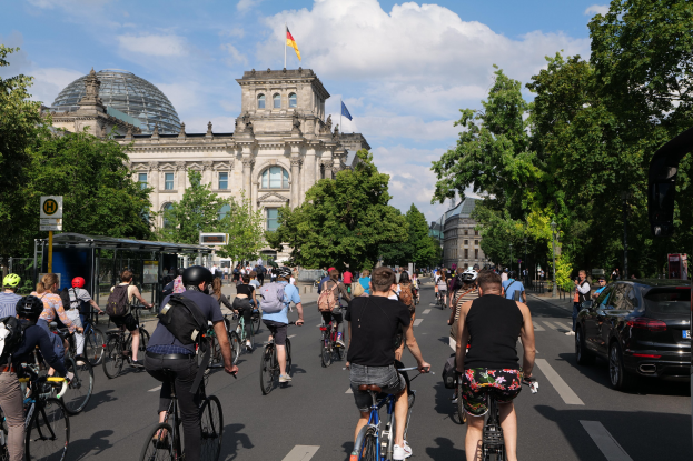 Gruppe von Menschen auf Fahrrädern auf einer von Bäumen gesäumten Straße in Berlin, Deutschland, mit Gebäuden und einer Bushaltestelle, unter einem bewölkten Himmel und einer Flagge auf einem Gebäude.